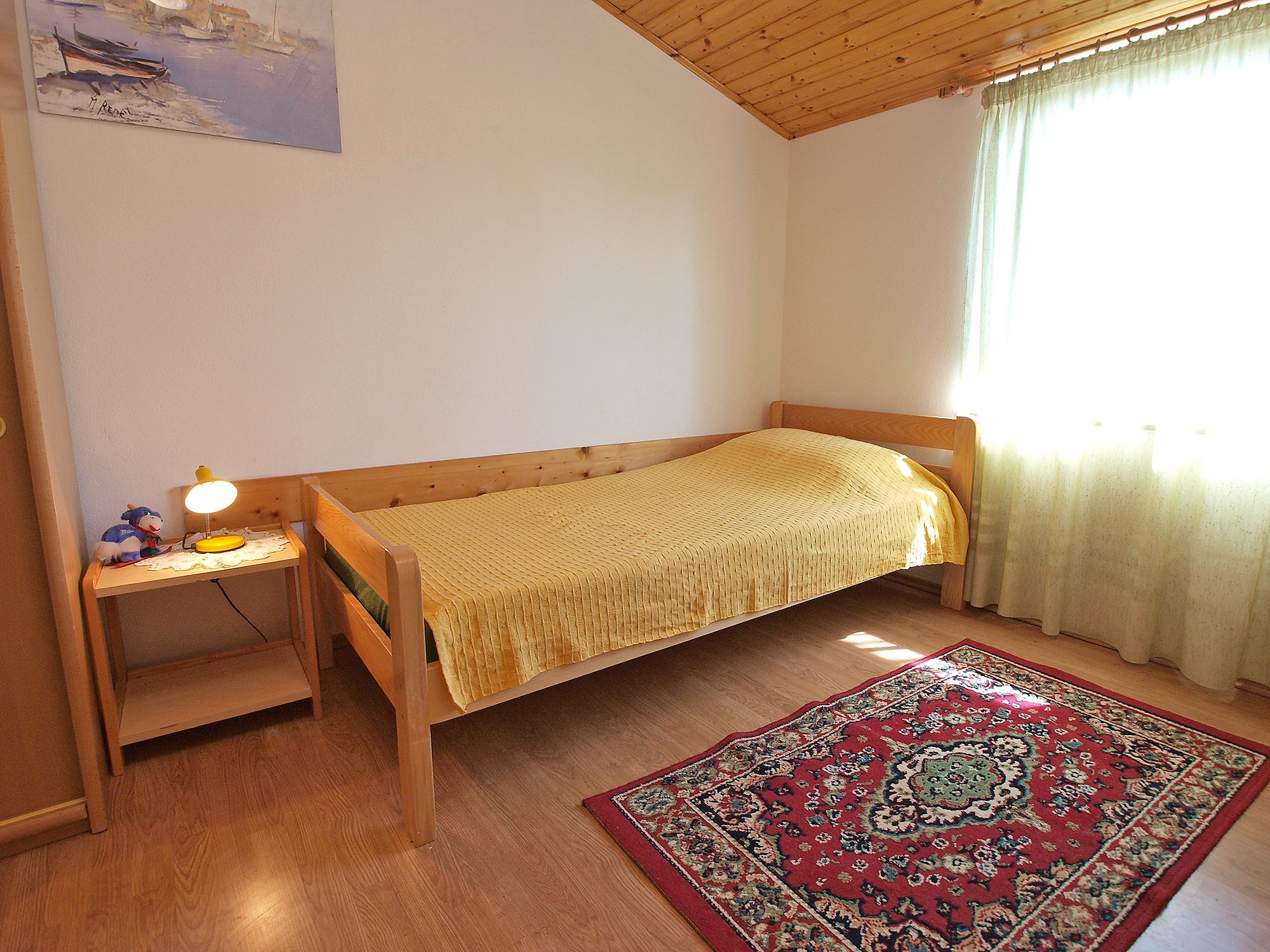 Quarto pequeno com uma cama de solteiro, cabeceira de madeira, mesa de cabeceira com abajur, tapete e um quadro na parede.
