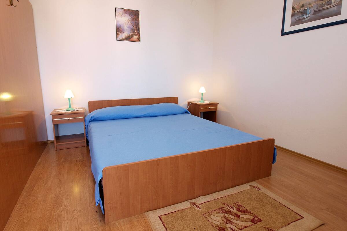 A bedroom with a wooden bed, two bedside tables with lamps, a small rug, and two framed pictures on the walls.