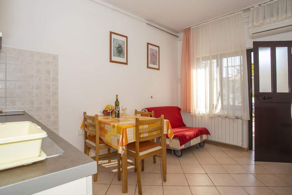 A compact kitchen with a dining table, chairs, and a small refrigerator. There is a window with curtains and a door to the right.