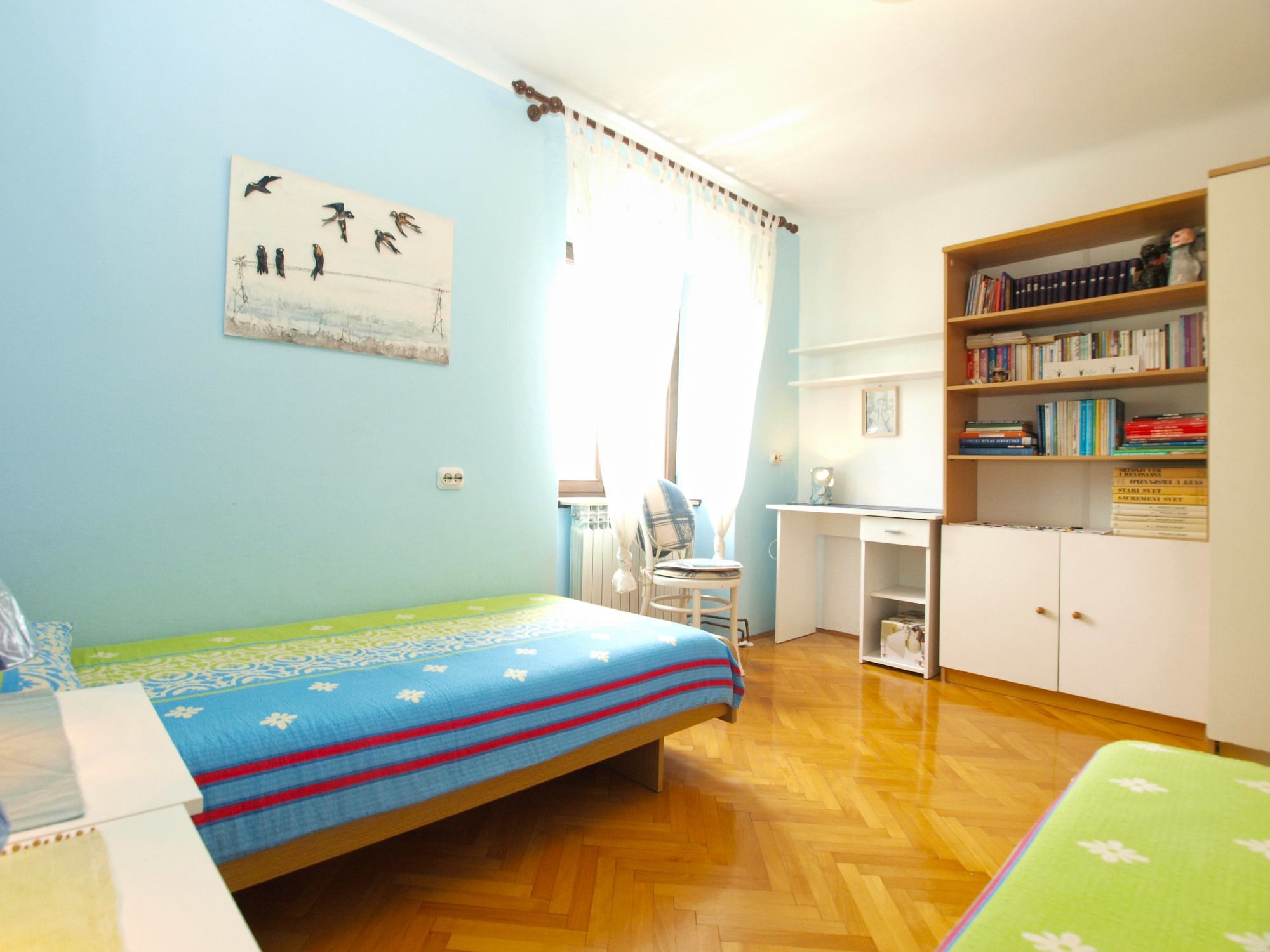 A bedroom with a bed, a desk, a chair, a bookshelf, and a window with curtains.