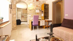 A living room with a sofa, a coffee table with items, a desk with a chair, and a kitchen in the background. - Apartment 64 (Vodnjan)