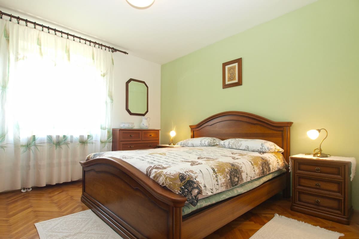 A bedroom with a wooden bed, bedside tables, a nightstand, a mirror, and a window with curtains.