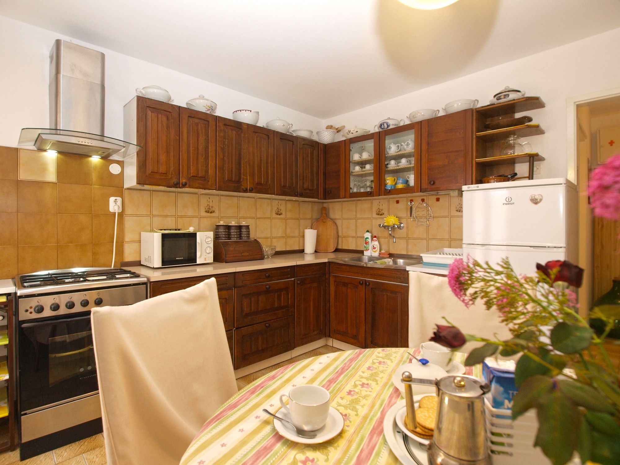 Een keuken met houten kastjes, een gasfornuis, een magnetron, een koelkast en een eettafel met een bloemrijk tafelkleed.