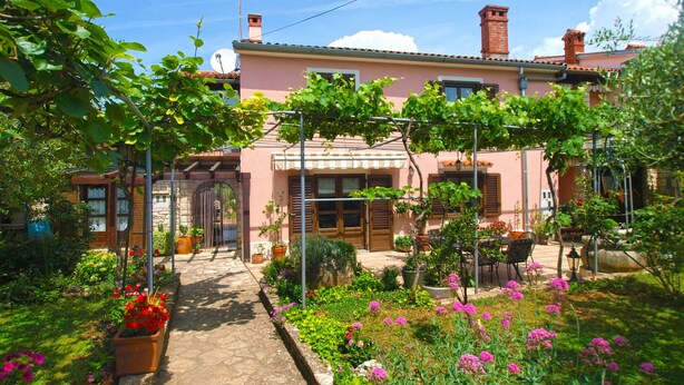 A pink villa with a stone pathway leading to a wooden door, surrounded by lush gardens and outdoor seating. - Apartment 64 (Vodnjan)