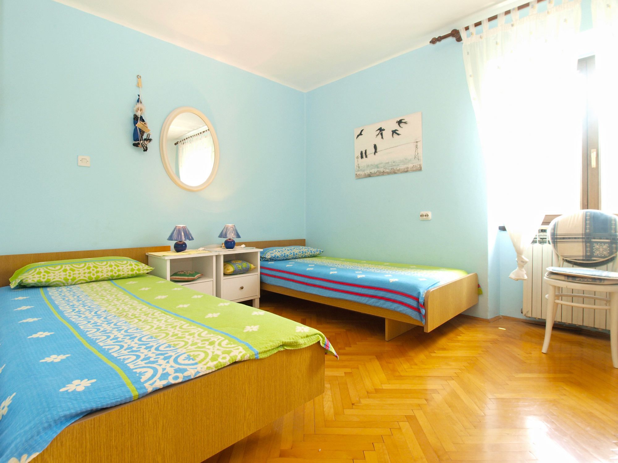 A bedroom with two single beds, a wooden floor, a desk with lamps, a chair, and a window with curtains.