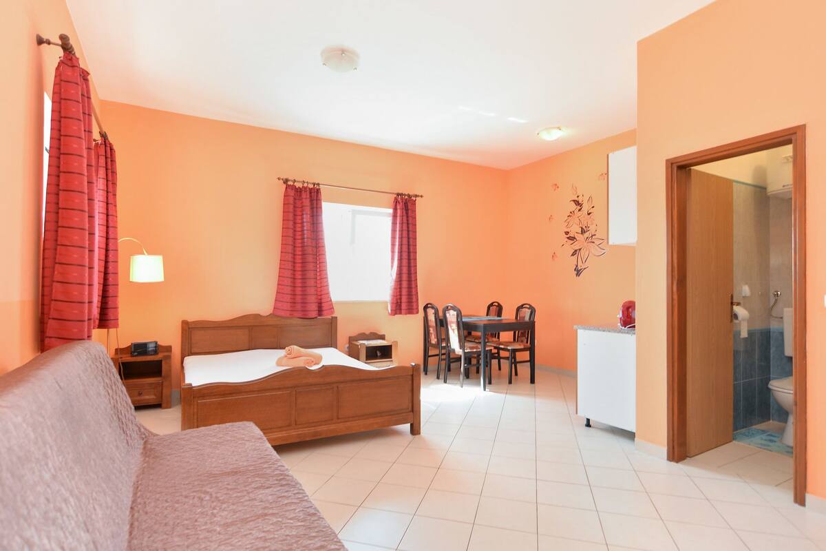 Une chambre à coucher avec un lit, une table de chevet, une lampe, une fenêtre avec des rideaux et une salle de bain visible à travers une porte ouverte.