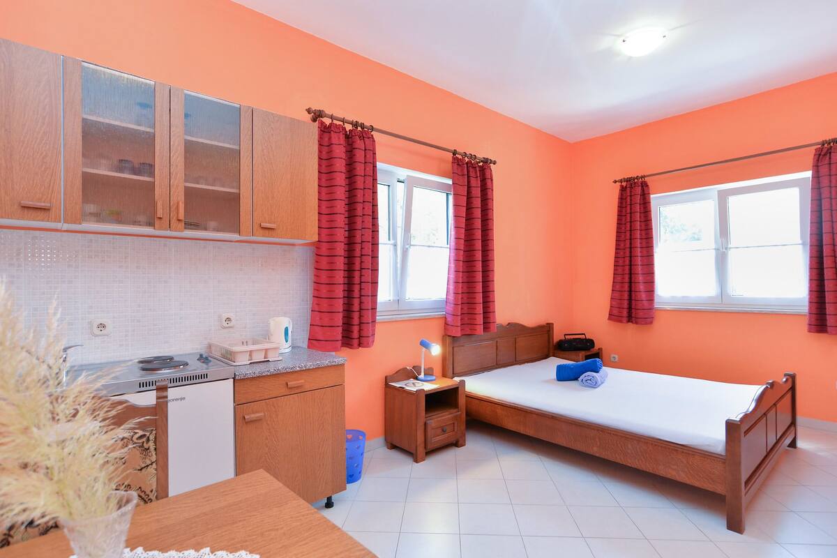 Une pièce de taille modeste comprenant un lit, une petite table de chevet, une commode, un coin cuisine et une fenêtre avec des rideaux.