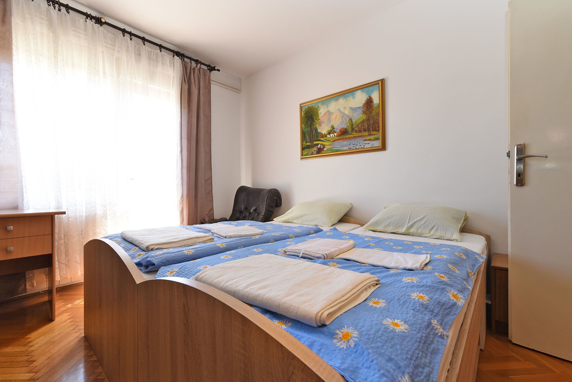 A bedroom with a wooden bed, a desk, and a chair. There is a painting on the wall and a door with a handle.