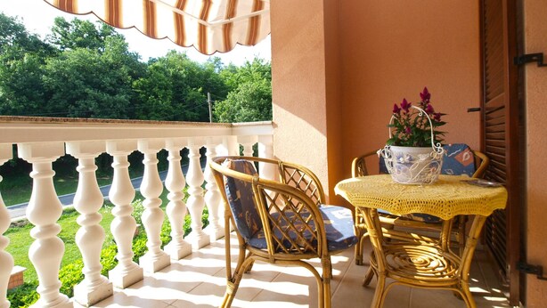A balcony with white balustrades, wicker chairs, a small table with a yellow tablecloth, and a potted plant with purple flowers. - Apartment 91 (Sveta Nedelja)