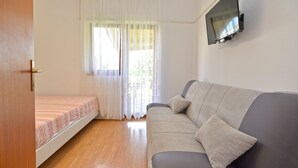 Una camera da letto con un letto, un divano grigio, una TV a muro e una finestra con tende leggere.