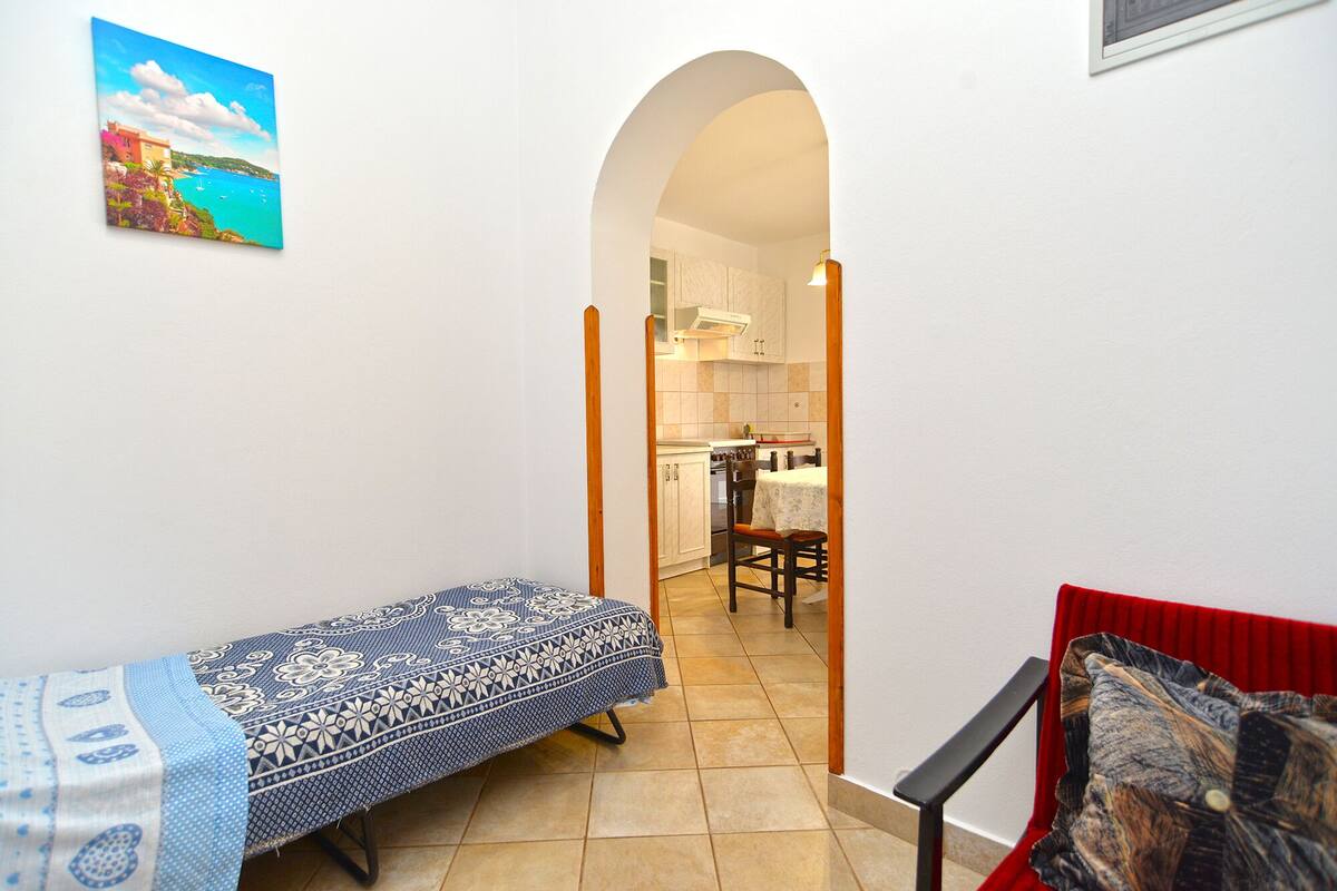 A narrow hallway with a bed on the left, a red chair on the right, and a kitchen in the background.