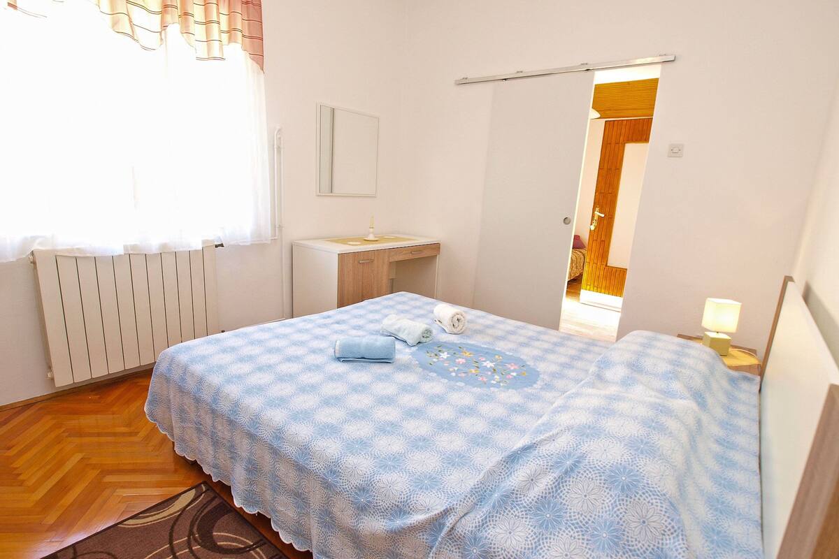 A bedroom with a bed, a bedside table, a mirror, a radiator, and a window with curtains.