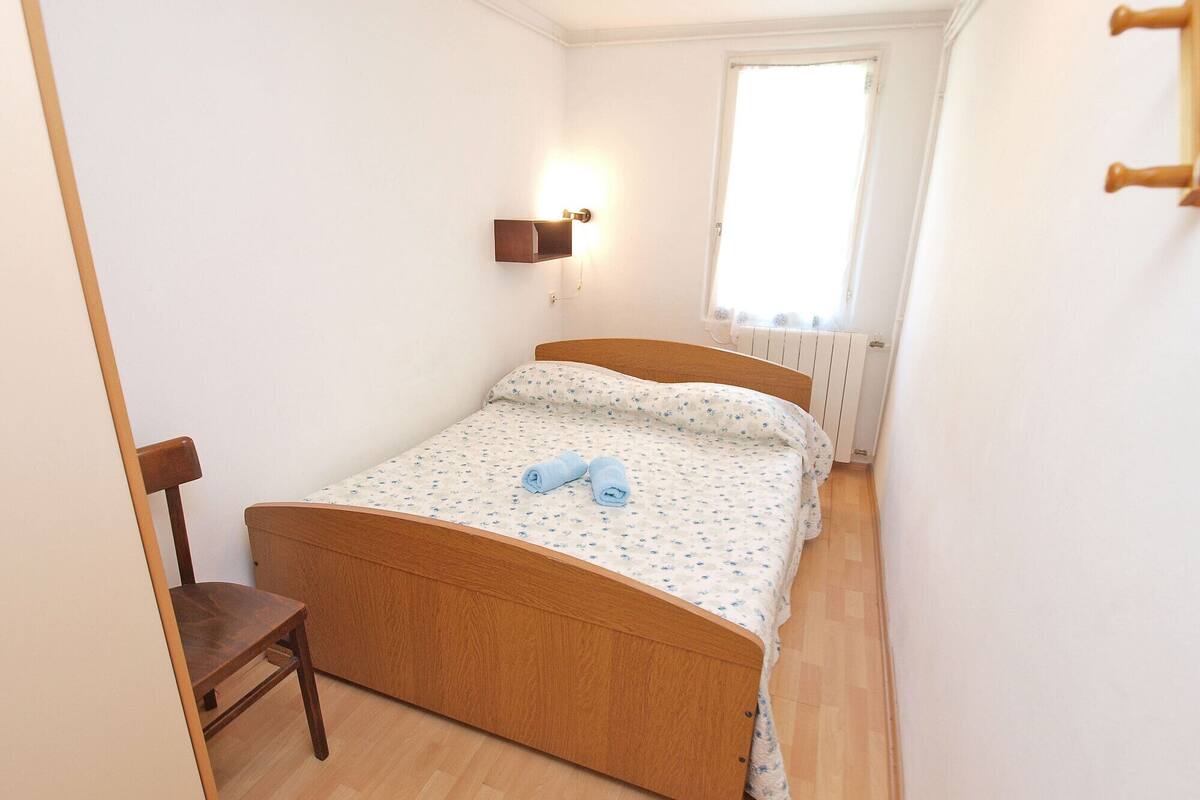 A small, neatly arranged bedroom with a wooden bed, a chair, a wall-mounted light, and a window with a view of the outside.