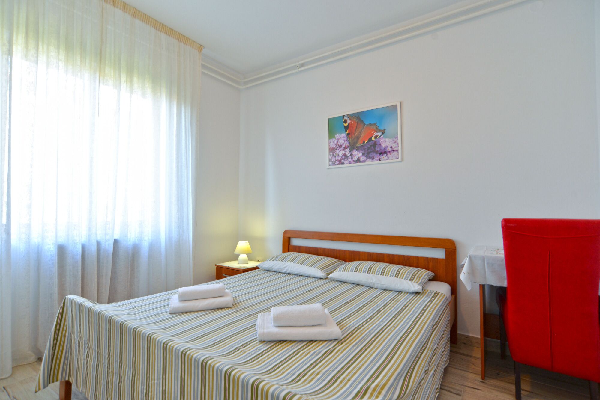 A hotel room with a bed, a nightstand, a red chair, and a painting on the wall.