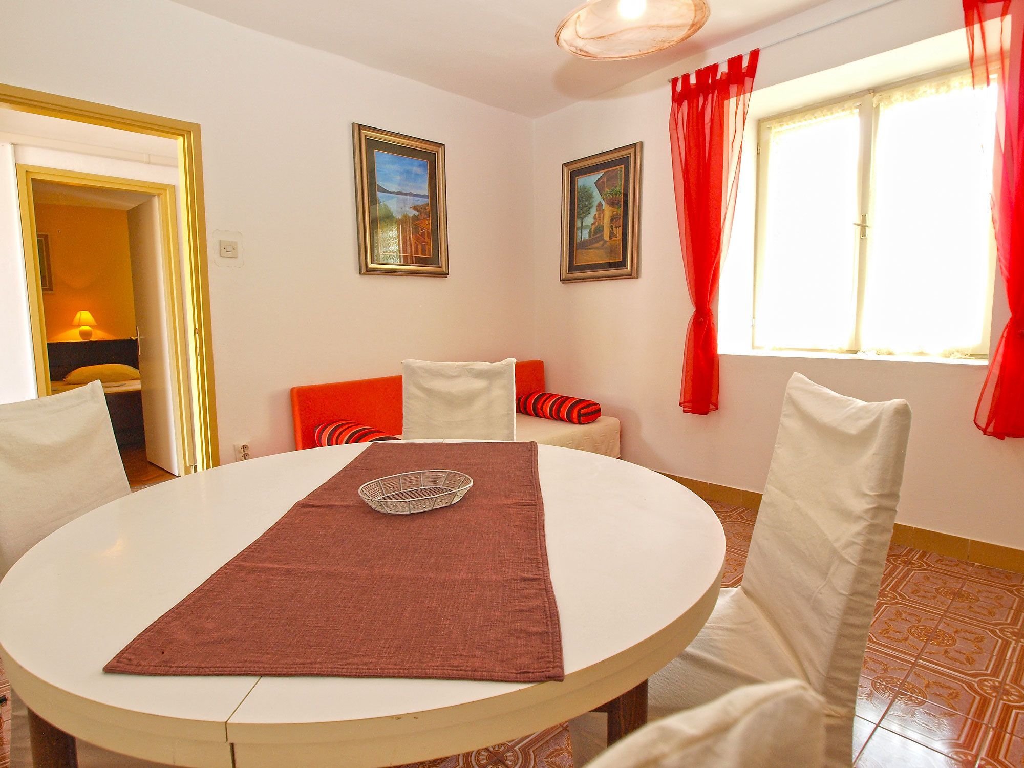 A dining area with a round table, chairs, and a red table runner. There are two framed pictures on the wall, and a window with red curtains.