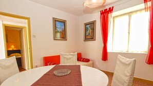 Un coin repas avec une table ronde, des chaises et un chemin de table rouge. Deux tableaux encadrés ornent le mur, et une fenêtre est agrémentée de rideaux rouges.