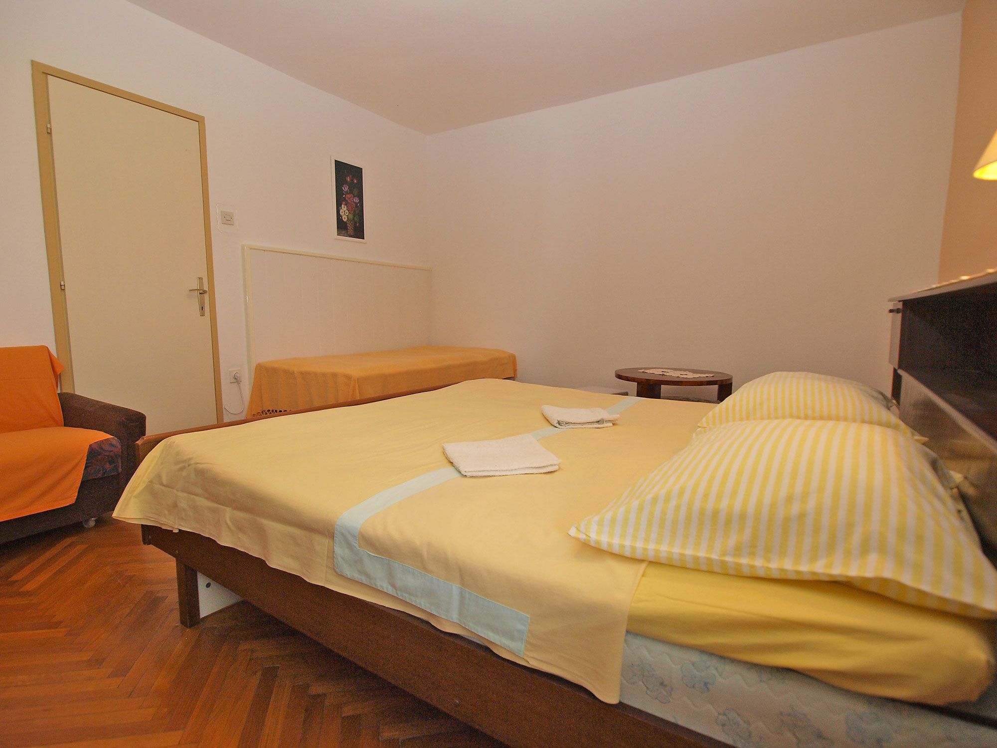 A single bed with a yellow bedspread, a bedside table, a chair, and a door.