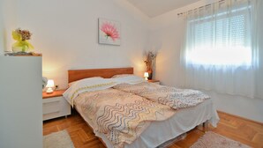 A bedroom with a wooden bed, white walls, a floral painting, a window with sheer curtains, and a bedside table with a lamp. - Apartment 258 (Pula)
