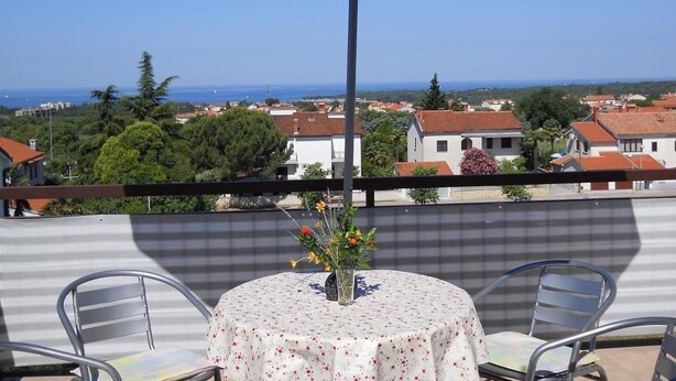 A balcony with a table and chairs, a blue umbrella, and a view of a coastal town. - Apartment 297 (Porec)