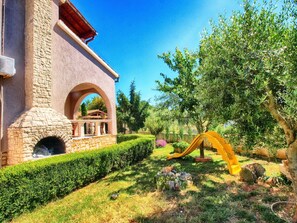 A stone building with a brick oven, a well-maintained garden with hedges, and a yellow slide.