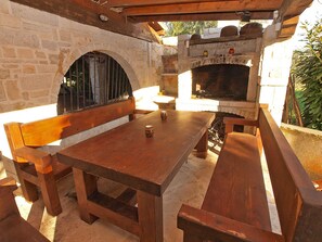 An outdoor patio with a stone wall, wooden table, benches, and a brick oven.