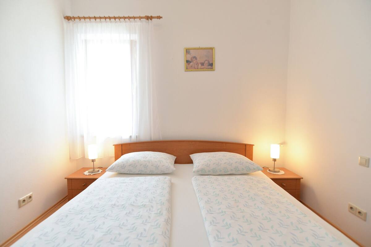 A double bed with a leaf-patterned bedspread, two bedside lamps, a wooden headboard, a window with sheer curtains, and a framed picture on the wall.