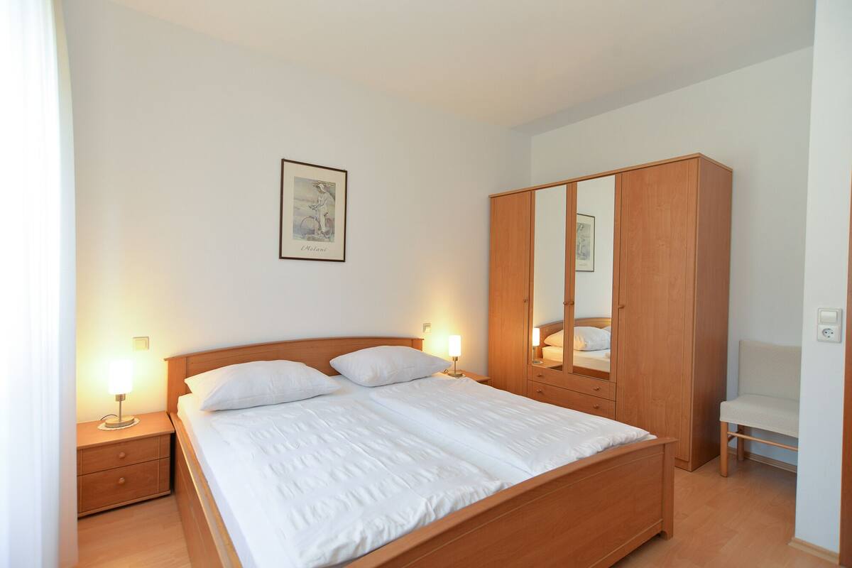 A neatly made bed with two pillows, a wooden wardrobe, a bedside table with a lamp, and a framed picture on the wall.