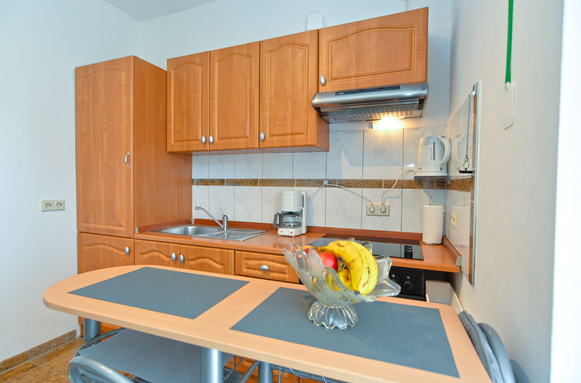 A compact kitchen with wooden cabinets, a built-in stove, a coffee maker, and a fruit bowl on the counter.