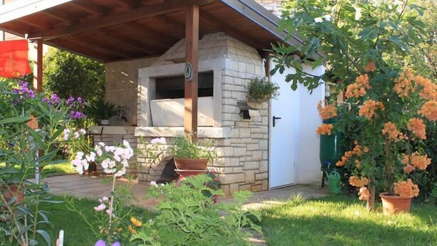 A stone-built outdoor kitchen with a chimney, surrounded by a garden with various plants and flowers. - Apartment 356 (Umag)