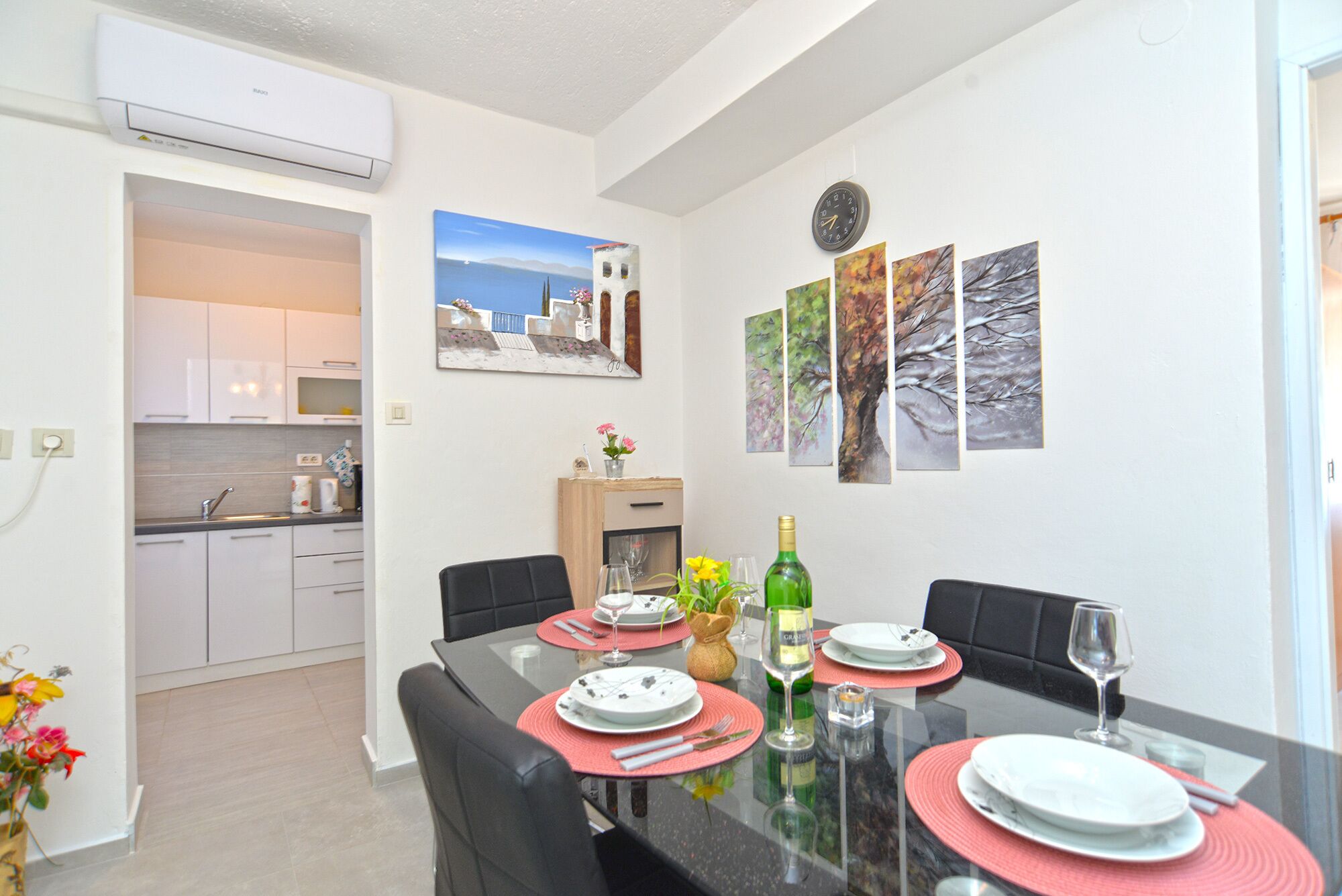 A dining area with a table set for four, a kitchen in the background, and artwork on the walls.