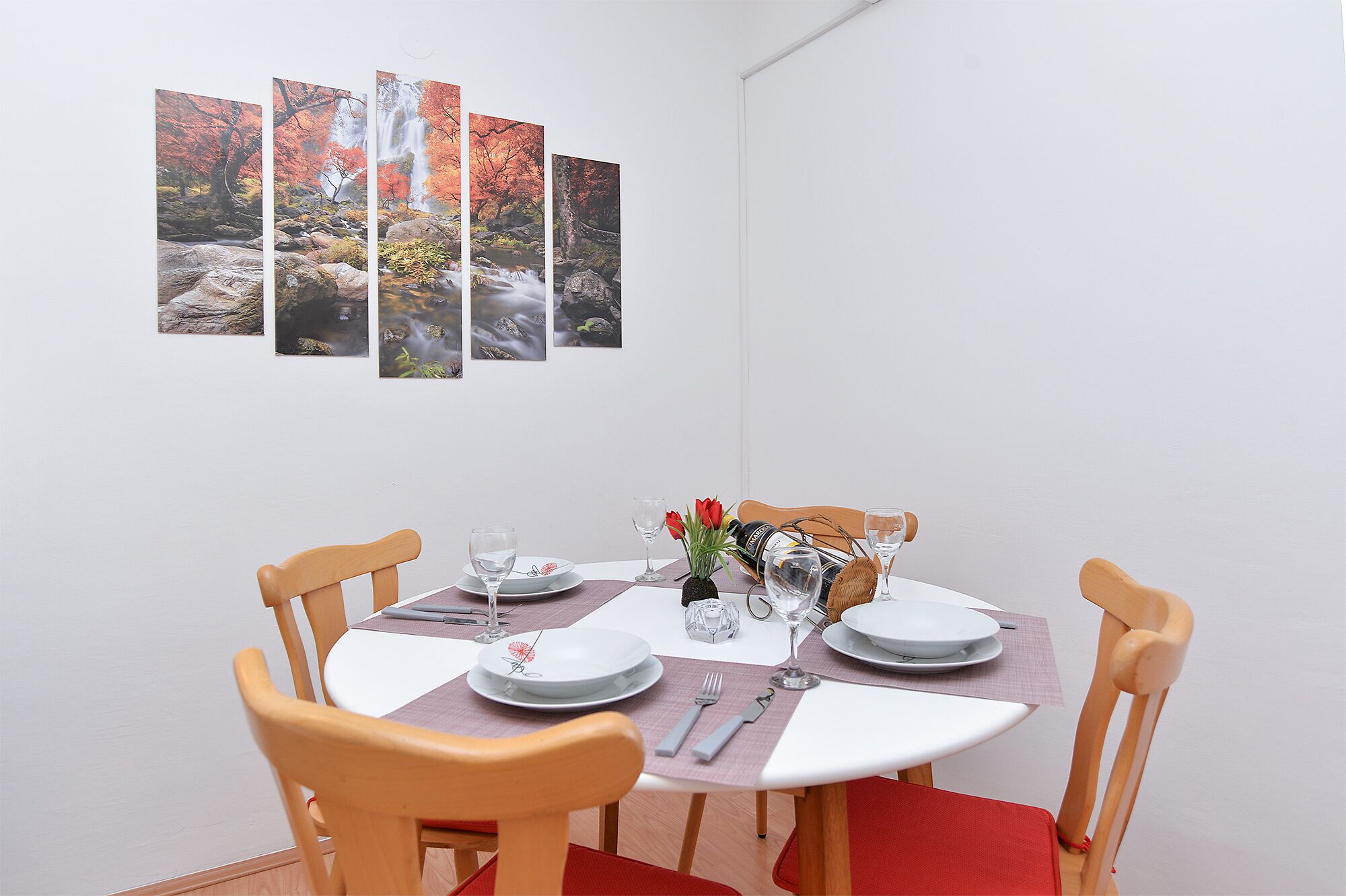 A dining area with a table set for two, featuring a white tablecloth, plates, cutlery, and a vase with red flowers.