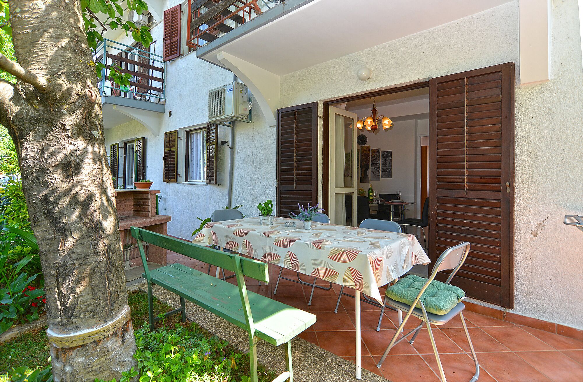 An outdoor patio with a table set for dining, surrounded by greenery and a tree.