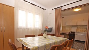 Ein Esszimmer mit einem Tisch, der für sechs Personen gedeckt ist, umgeben von Holzstühlen. Ein Fernseher ist an der Wand befestigt und im Hintergrund ist eine Küche zu sehen.