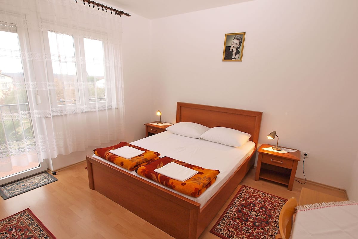 Une chambre à coucher bien aménagée, avec un lit en bois, deux tables de chevet dotées de lampes et une vue sur l’extérieur à travers une fenêtre ornée de rideaux légers.