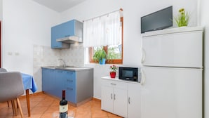 Een kleine keuken met blauwe kastjes, een witte koelkast, een magnetron, en een eethoek met een tafel en stoelen.