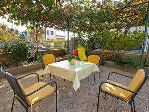 An outdoor patio with a table and four chairs, surrounded by greenery and a building in the background. - House 385 (Pula)