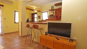 A compact living space with a kitchenette, a dining area with two stools, a TV on a wooden cabinet, and a large window providing natural light. - Apartment 389 (Pula)