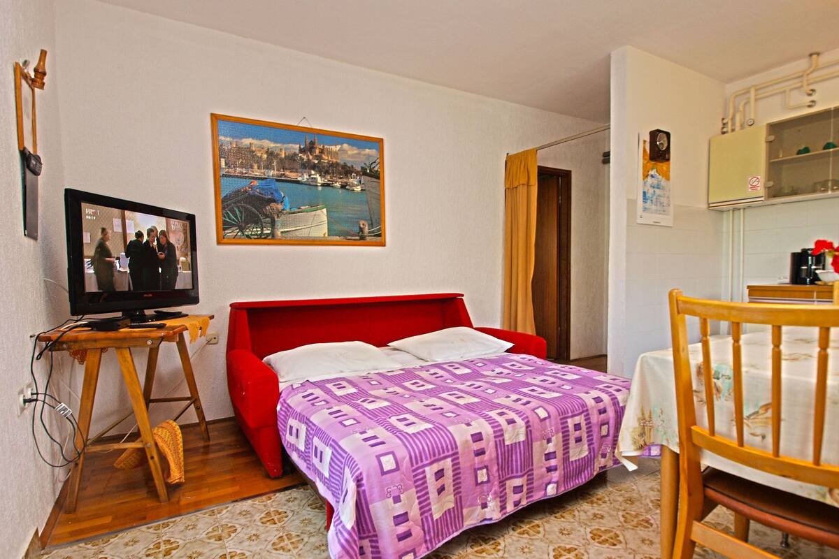 A room with a red bed, a television on a wooden stand, a painting of a coastal town, and a small table with a chair.