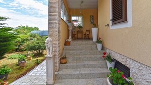 Una escalera de piedra que lleva a una puerta con ventanal, flanqueada por macetas con plantas y una estatua.
