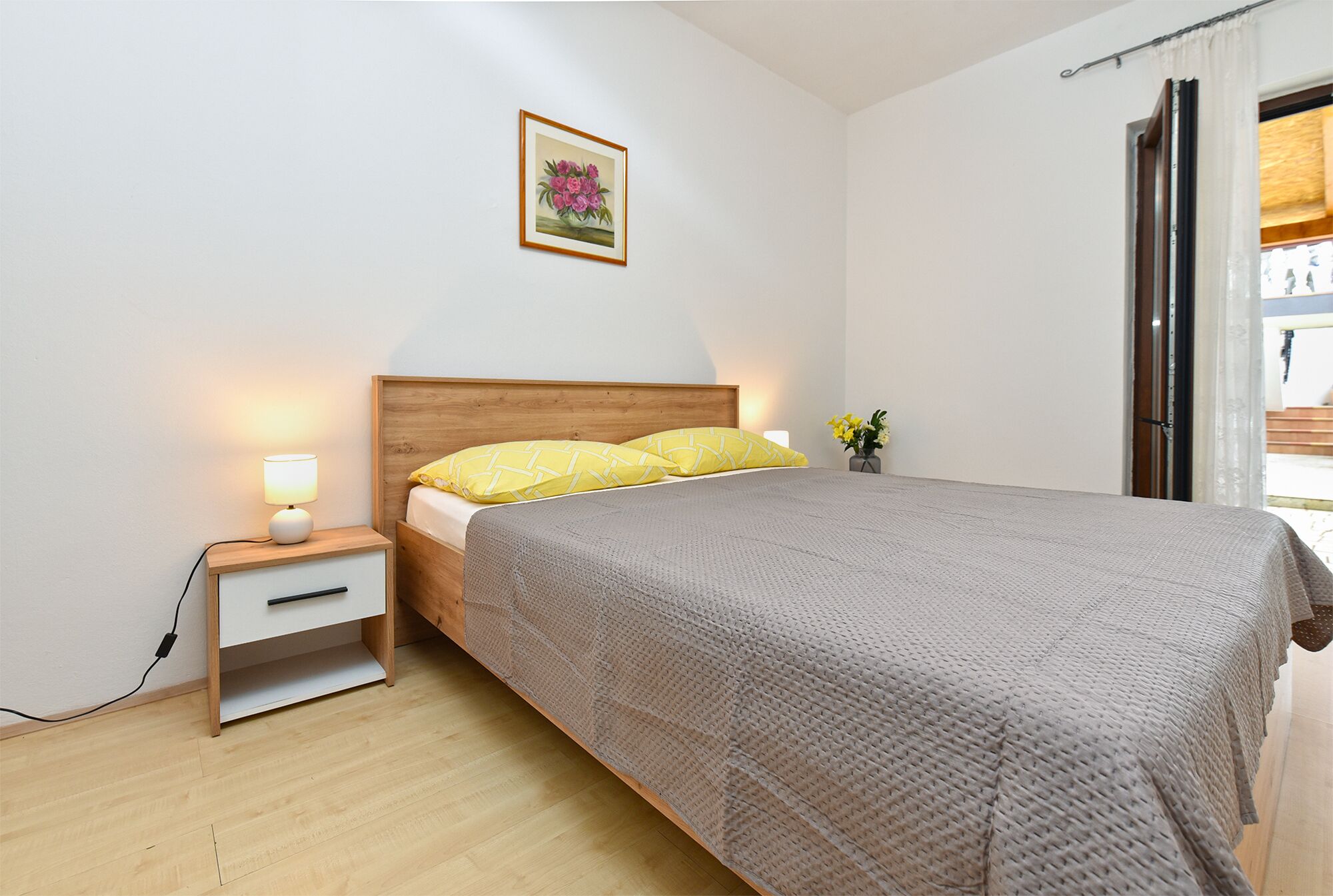 A bedroom with a wooden headboard, a bedside table with a lamp, a bed with a grey quilt, and a framed picture on the wall.