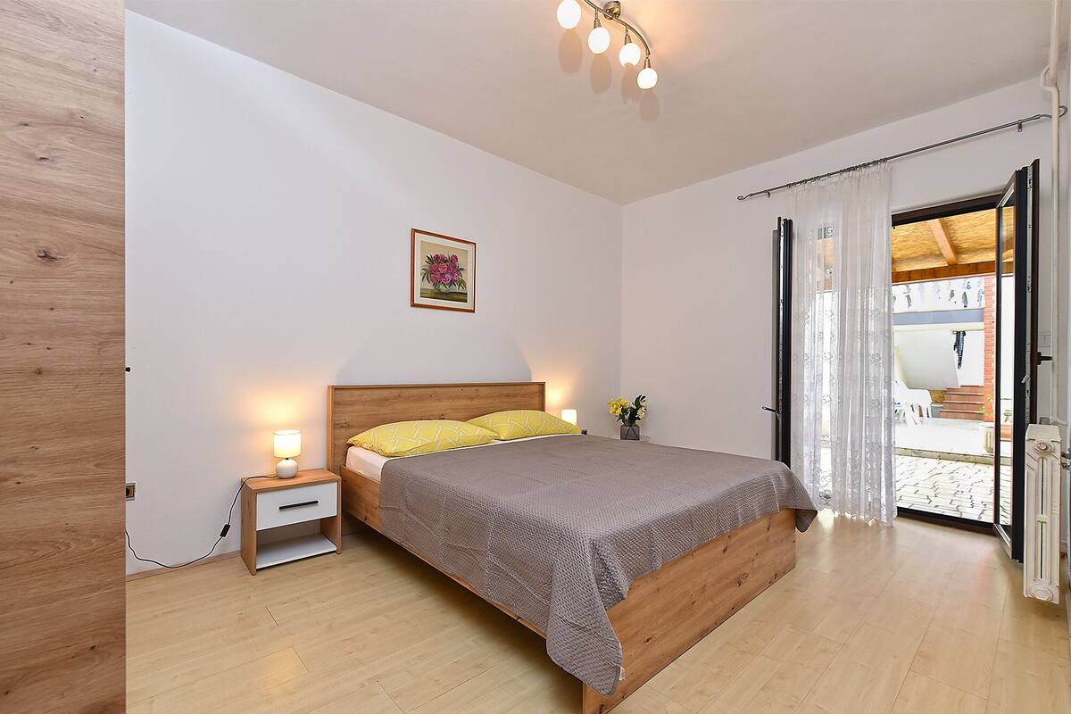 A bedroom with a wooden bed, a nightstand, a bedside lamp, a framed picture on the wall, and a view of the outside through a door.