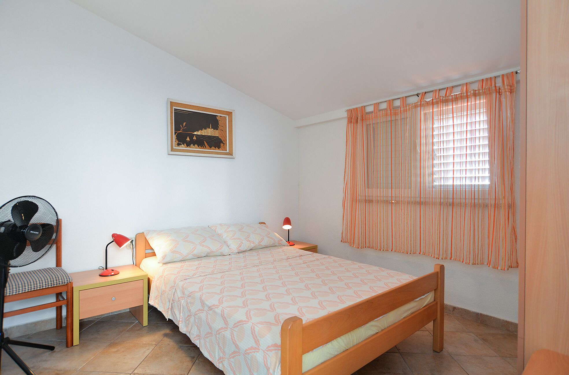 Una camera da letto con un letto, una scrivania, una sedia, una ventola, una lampada e una finestra con tende.