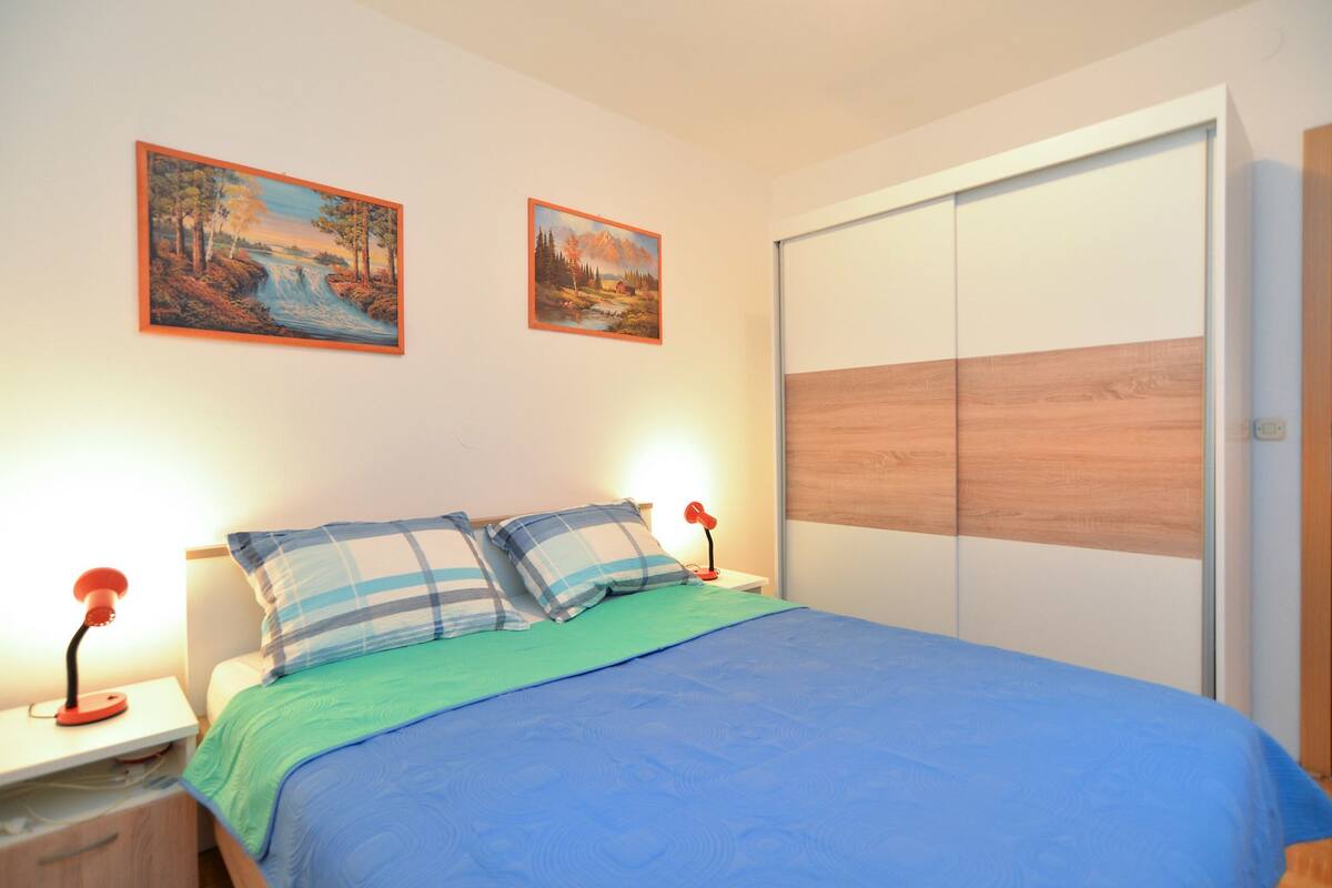 A bedroom with a bed, two nightstands, a red lamp, and two framed landscape paintings on the wall.
