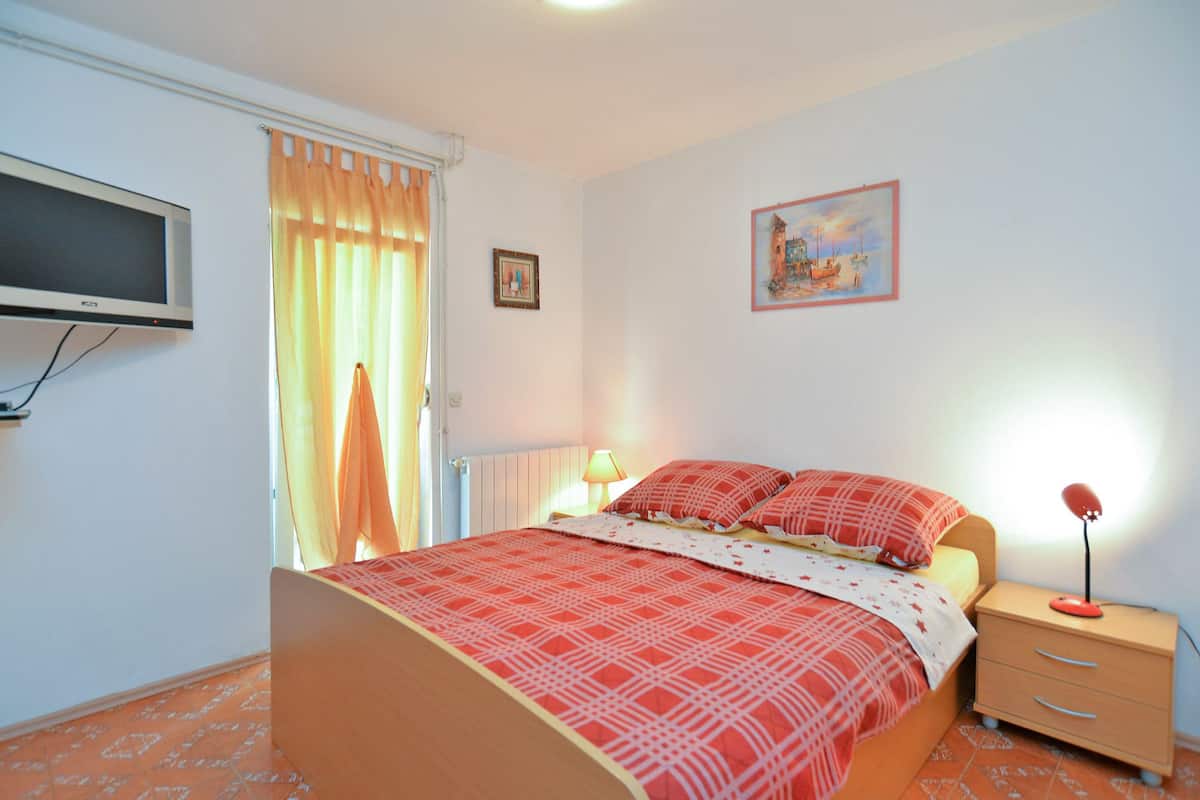 A bedroom with a bed, a bedside table, a lamp, a TV mounted on the wall, and a window with curtains.