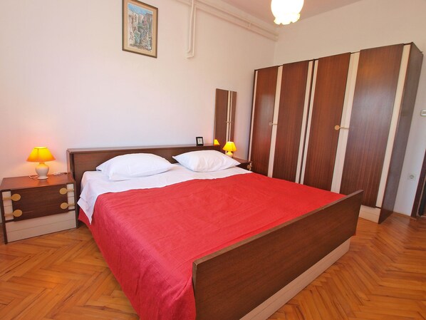 Un lit bien fait, avec des oreillers blancs et une couvre-lit rouge, dans une pièce au plancher en bois. On y trouve deux tables de chevet avec des lampes, ainsi qu’une armoire à rayures verticales.