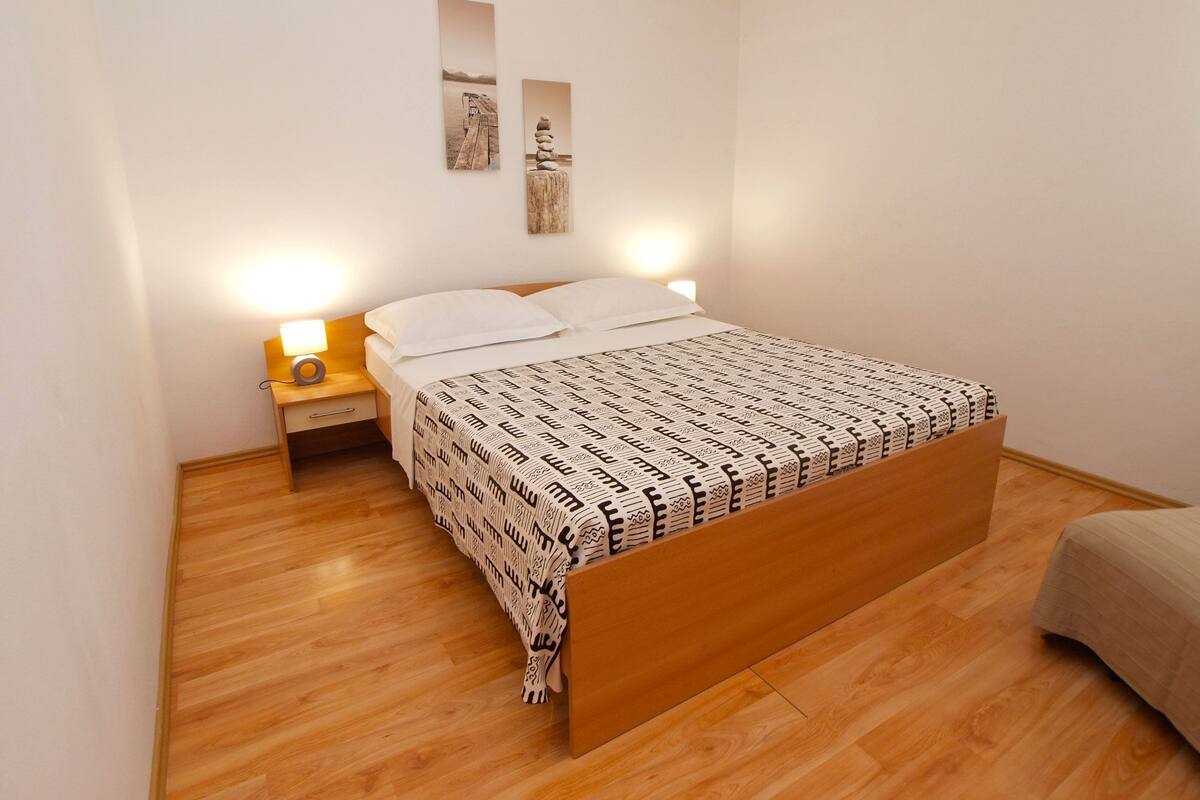 Un dormitorio con cama de madera, ropa de cama blanca, dos lámparas de pared y una mesita de noche de madera con lámpara.