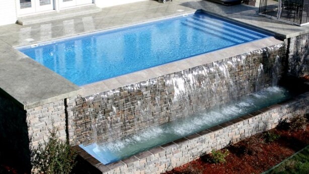 Een rechthoekig zwembad met een waterval, omringd door een stenen muur en een verhard terras.