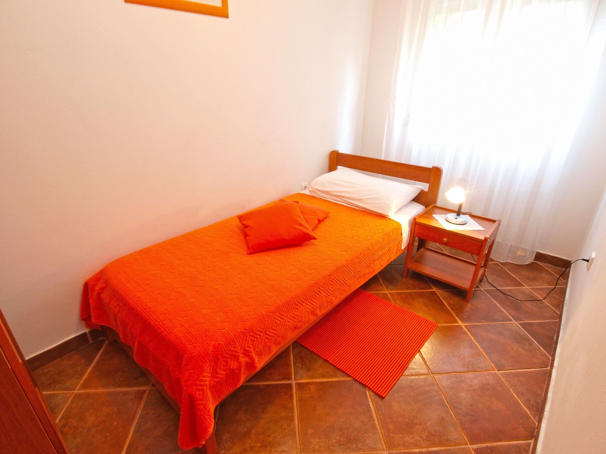 A single bed with an orange bedspread and pillows, a bedside table with a lamp, and a red rug on a tiled floor.