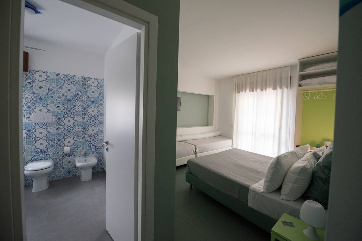 A bedroom with a bed, a bedside table, a nightstand, a window with curtains, and a bathroom with a toilet and sink.