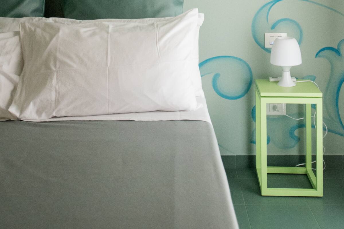A neatly made bed with white linens and a green pillow, a small green nightstand with a white lamp, and a wall with blue floral patterns.
